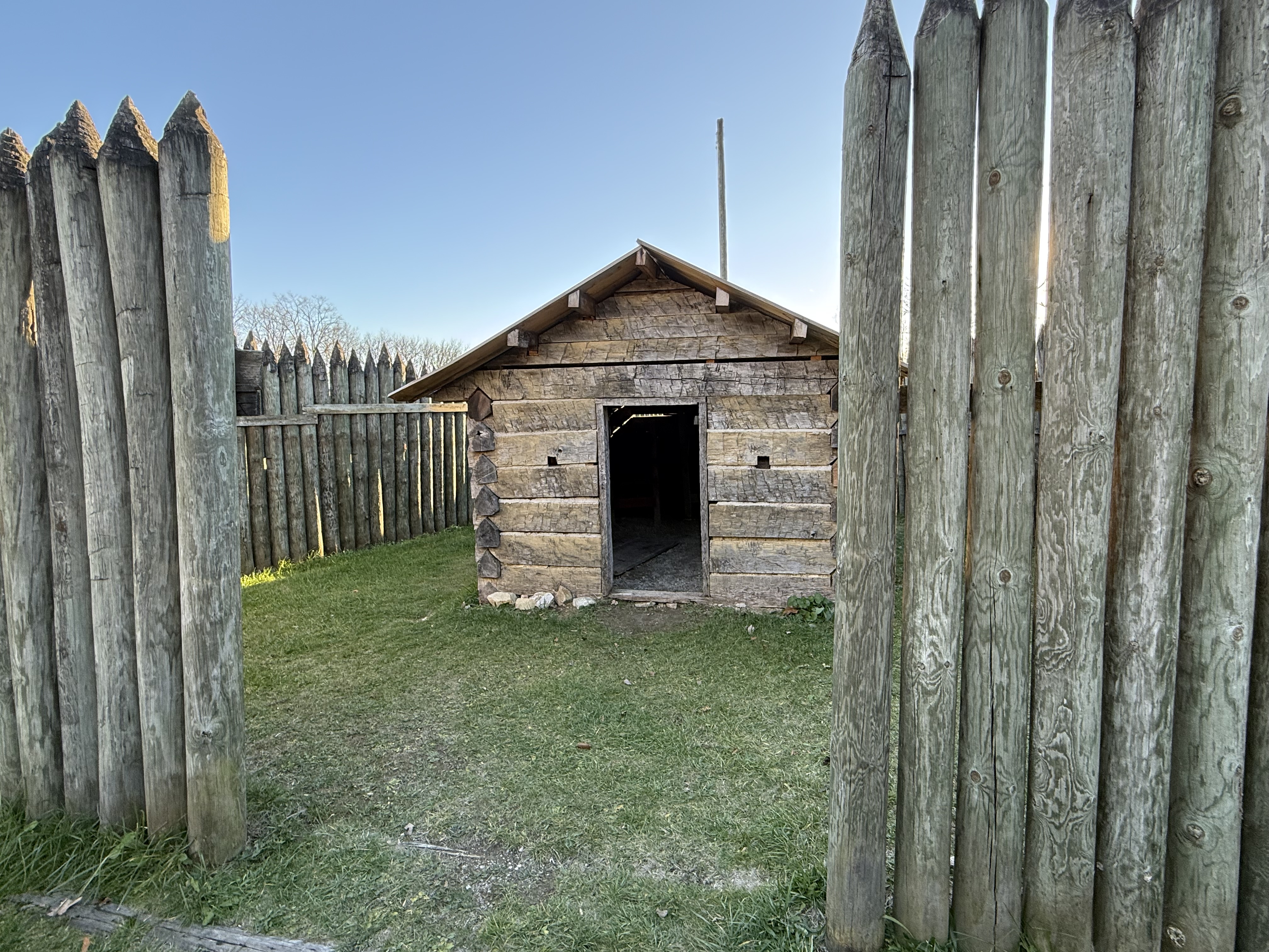 A view from inside the fort, looking through a palisade gate at a small log cabin.